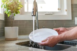 two hands holding white dish under running kitchen faucet rinsing plate